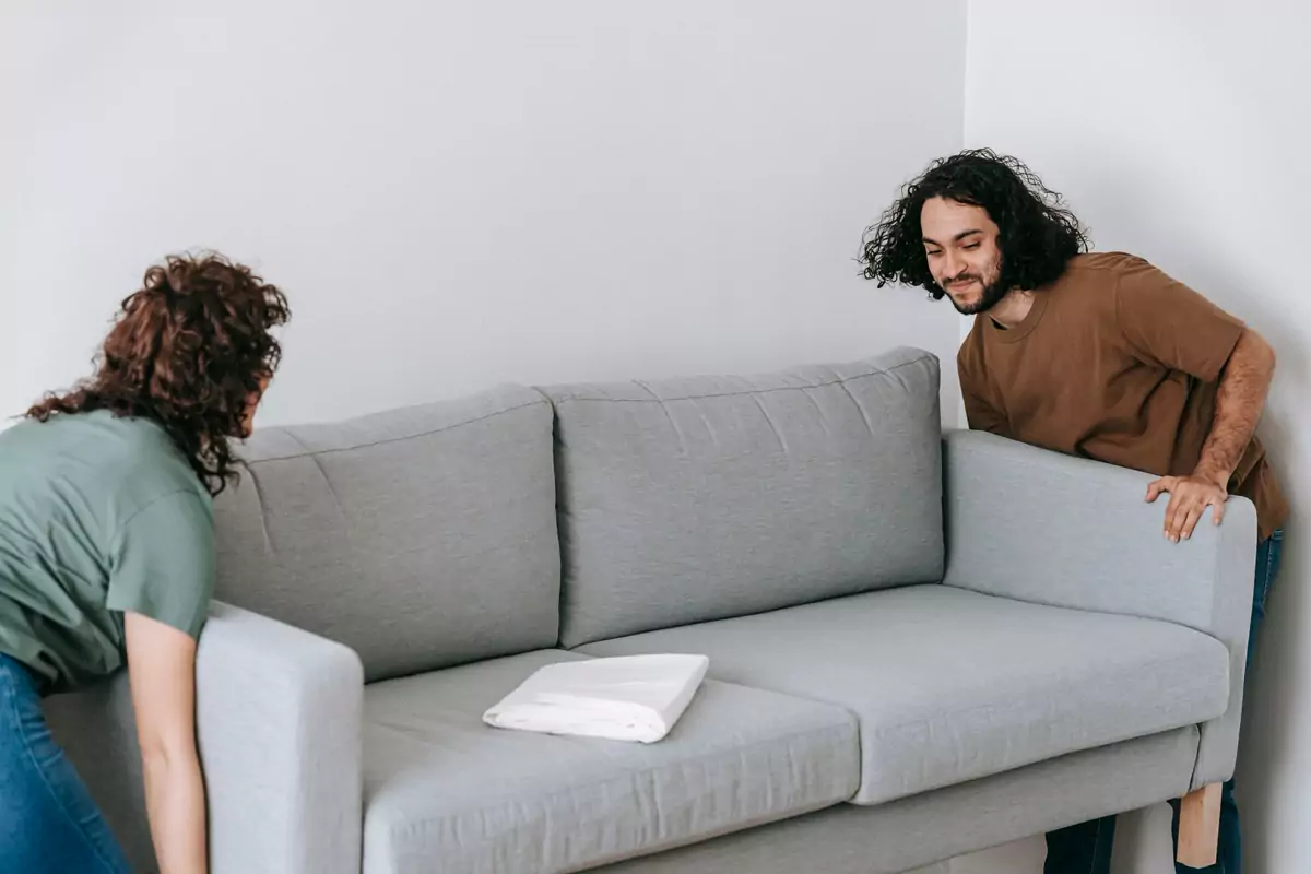 Zwei Personen tragen gemeinsam ein Sofa durch ein helles Wohnzimmer